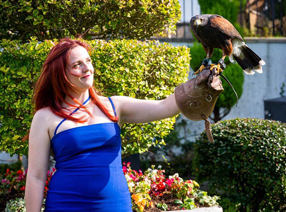 young woman in blue dress holding a hawk on her arm in a garden with flowers and greenery featuring two birds and nature